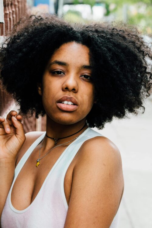 Person with curly hair wears a white tank top and looks at the camera
